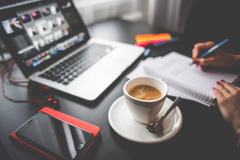 Laptop, coffee cup and phone on a desk while a person writes notes