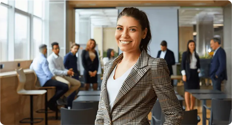 Smiling businesswoman in a checked blazer standing in a busy open-plan office with colleagues behind her
