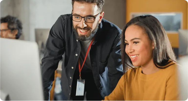 Smiling man with glasses and an ID lanyard leaning over a colleague's desk as she works at her computer