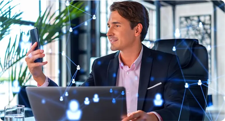 Businessman at a laptop holding his phone, with glowing network connection icons linking people across the office
