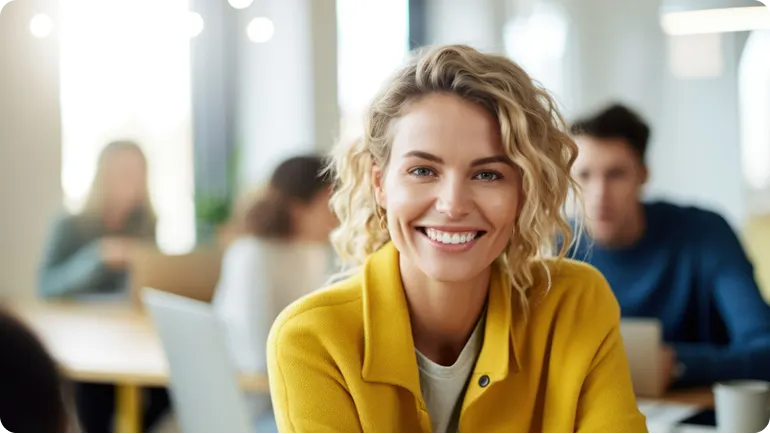 Smiling woman with curly blonde hair in a yellow jacket sitting in a bright office with colleagues behind her