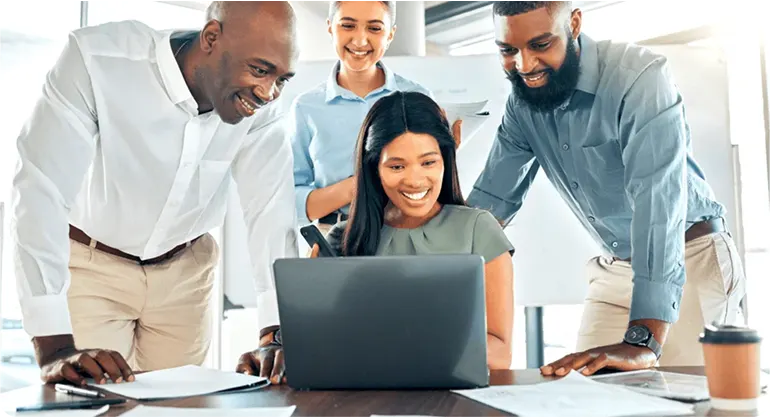 Team of four colleagues gathered around a laptop at a desk, smiling as they review work together